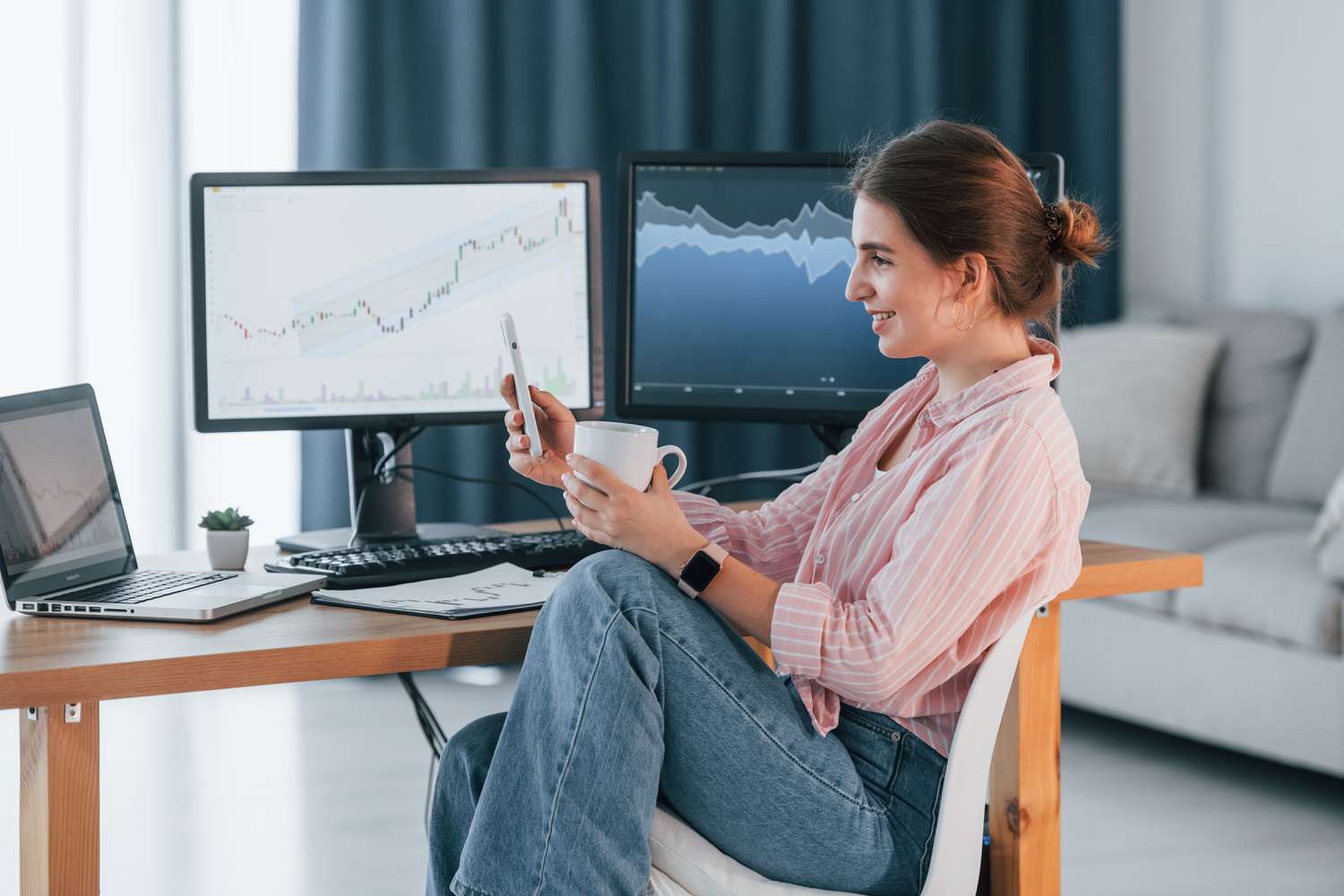 Woman holding a cup and her phone sitting in front of two desktops