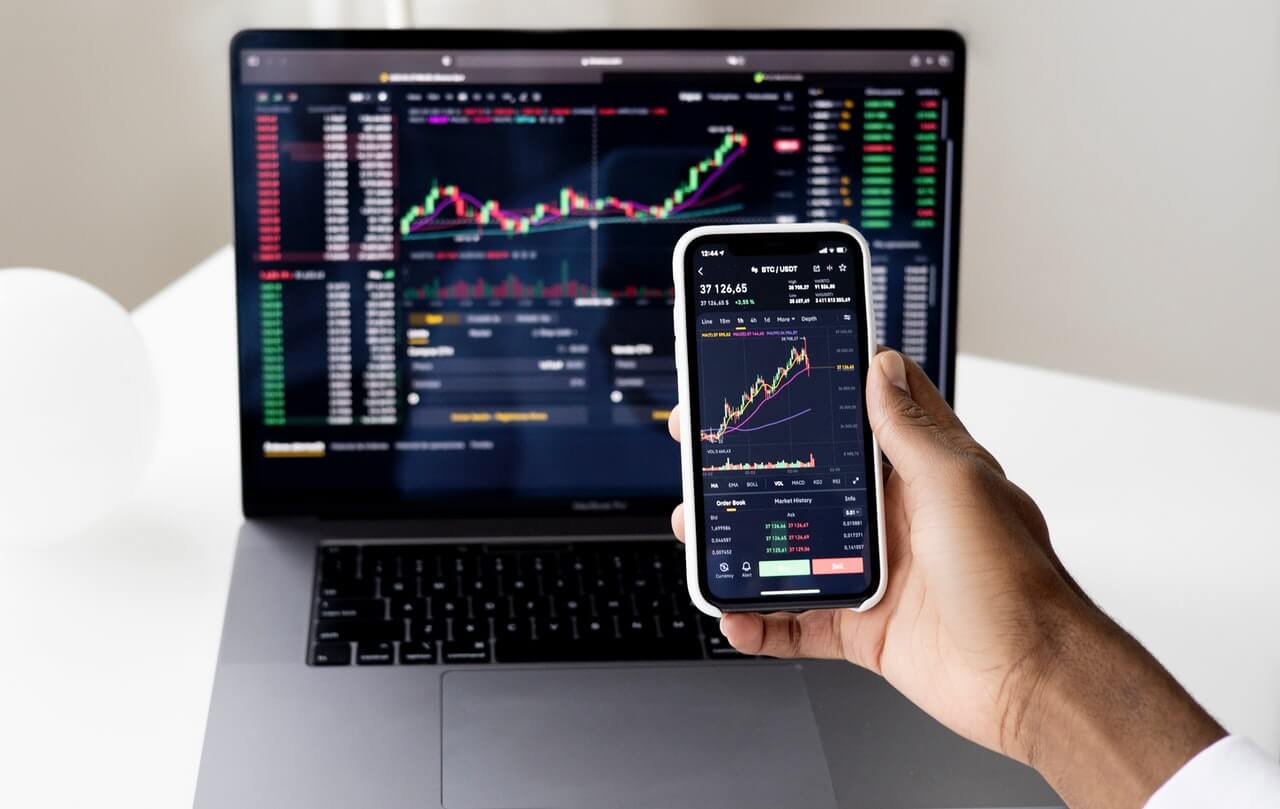 A person holding a smartphone showing a cryptocurrency trading chart in front of a laptop with the same chart.